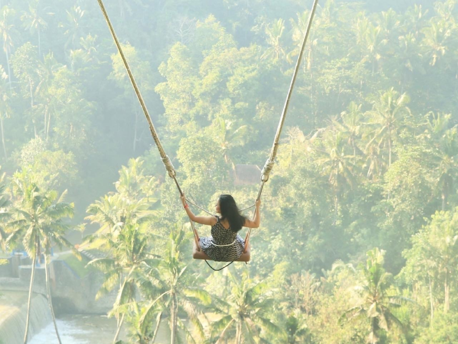 A woman in a long red dress swinging high above a lush green jungle valley on the famous Bali Swing.